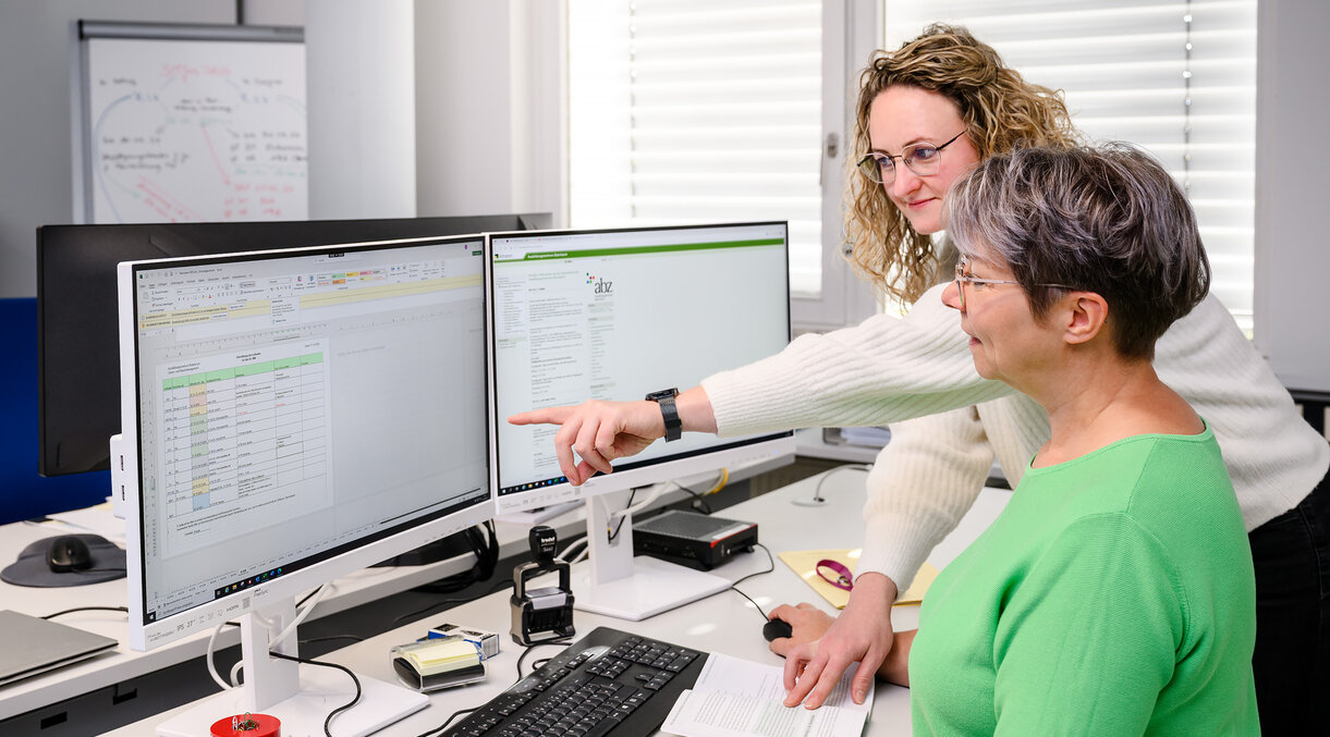 Zwei Sachbearbeiterinnen beraten sich an einem Schreibtisch; eine zeigt etwas auf einem der beiden Monitore