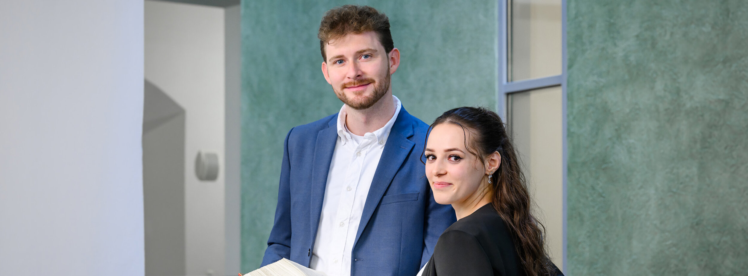 Ein Rechtspfleger und eine Rechtspflegerin stehen im Gang; der Rechtspfleger hält ein aufgeklapptes Buch