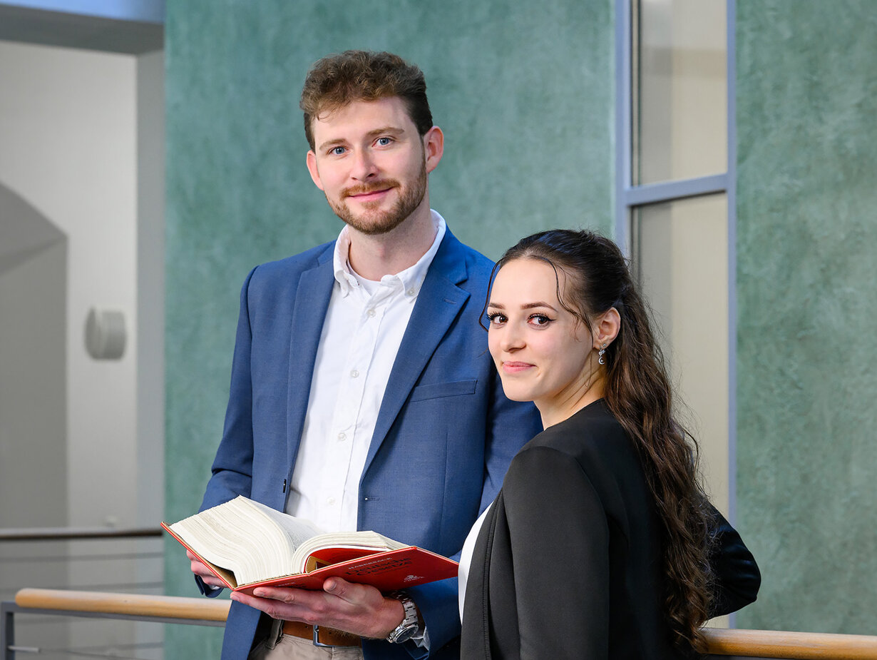 Ein Rechtspfleger und eine Rechtspflegerin stehen im Gang; der Rechtspfleger hält ein aufgeklapptes Buch