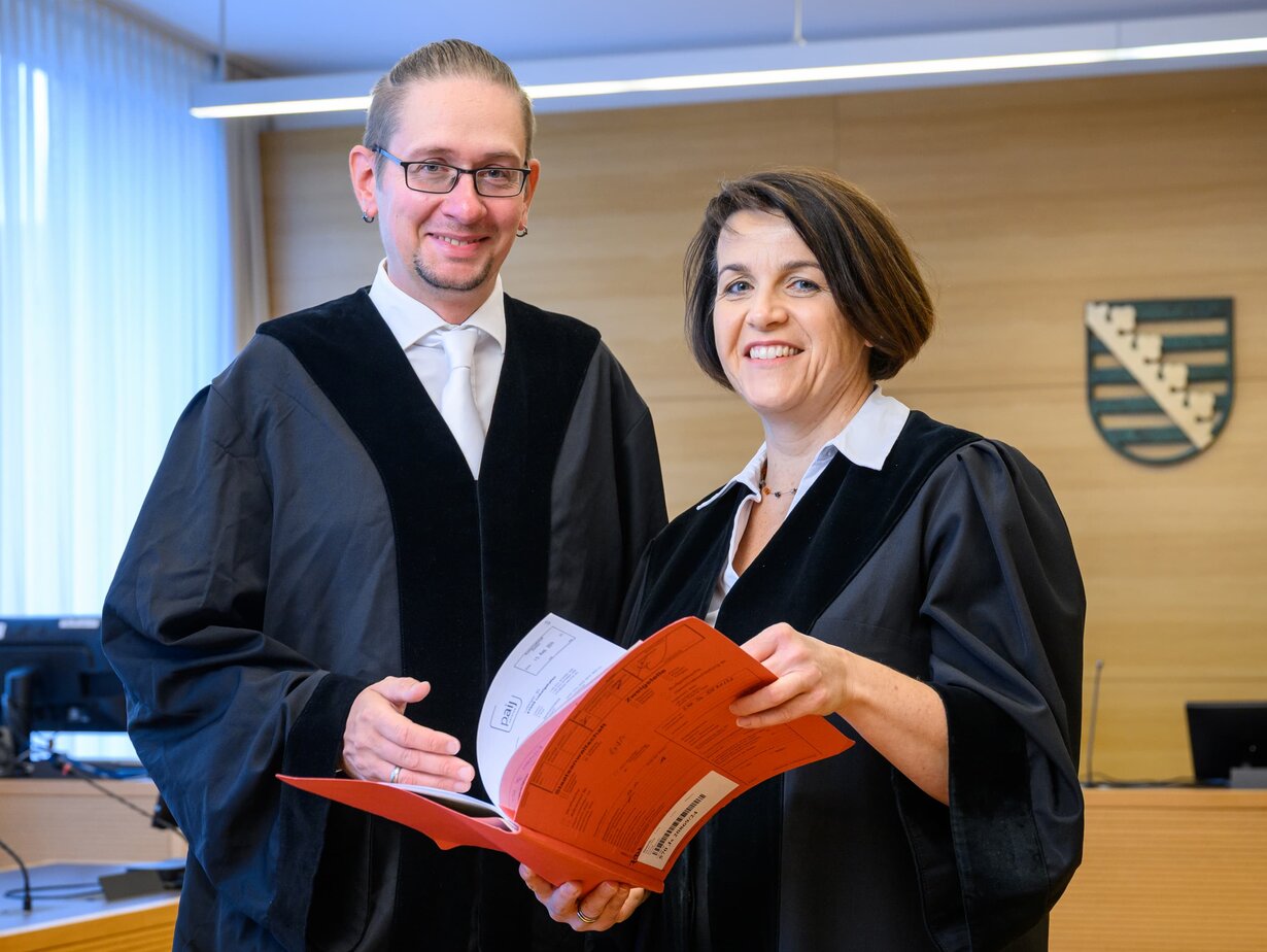 Ein Amtsanwalt und eine Amtsanwältin in einem Gerichtssaal und halten eine Akte geöffnet vor sich