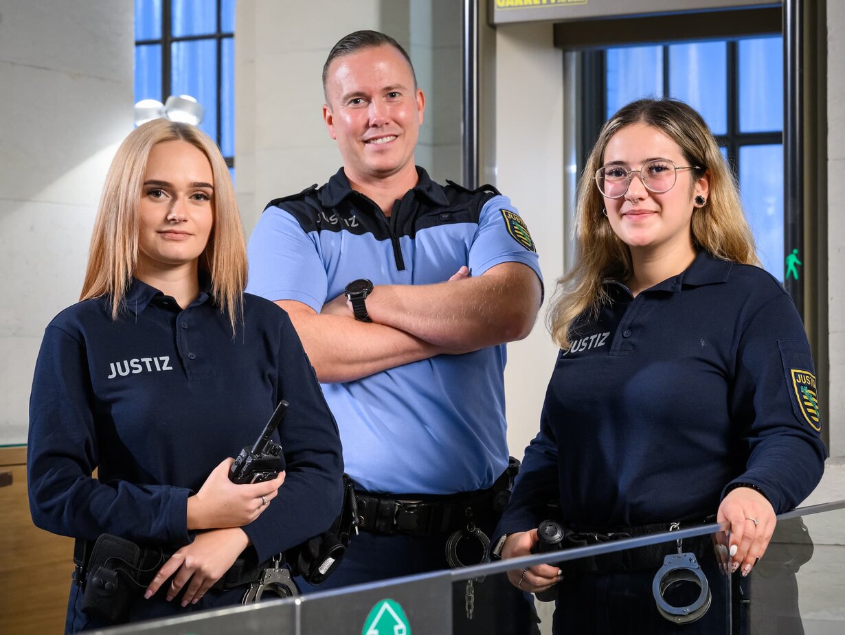 Eine Gruppe von Justizwachtmeistern im Gerichtsgebäude; der Justizwachtmeister in der Mitte hat die Arme verschränkt vor dem Körper