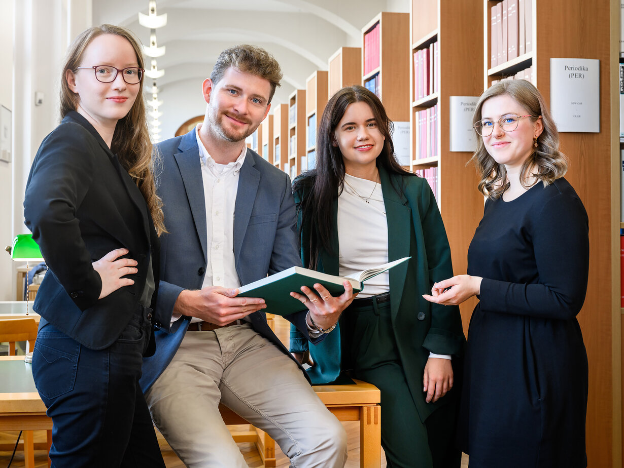 Eine Gruppe von Rechtspflegern in der Bibliothek; ein Rechtspfleger in der Mitte hält ein aufgeklapptes Buch