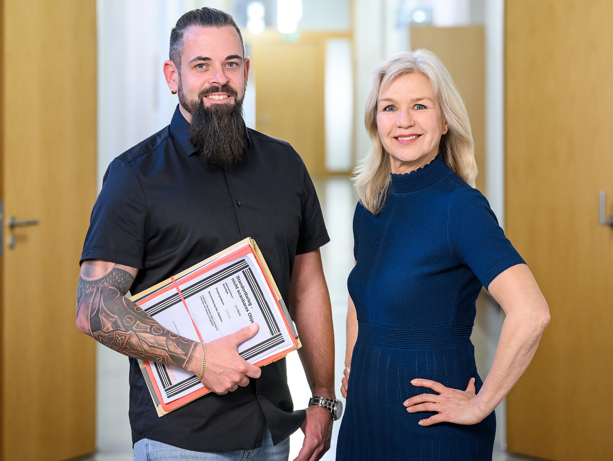 Ein Justizsekretär und eine Justizsekretärin vor dem Büro; der Justizsekretär hält eine Akte in der Hand