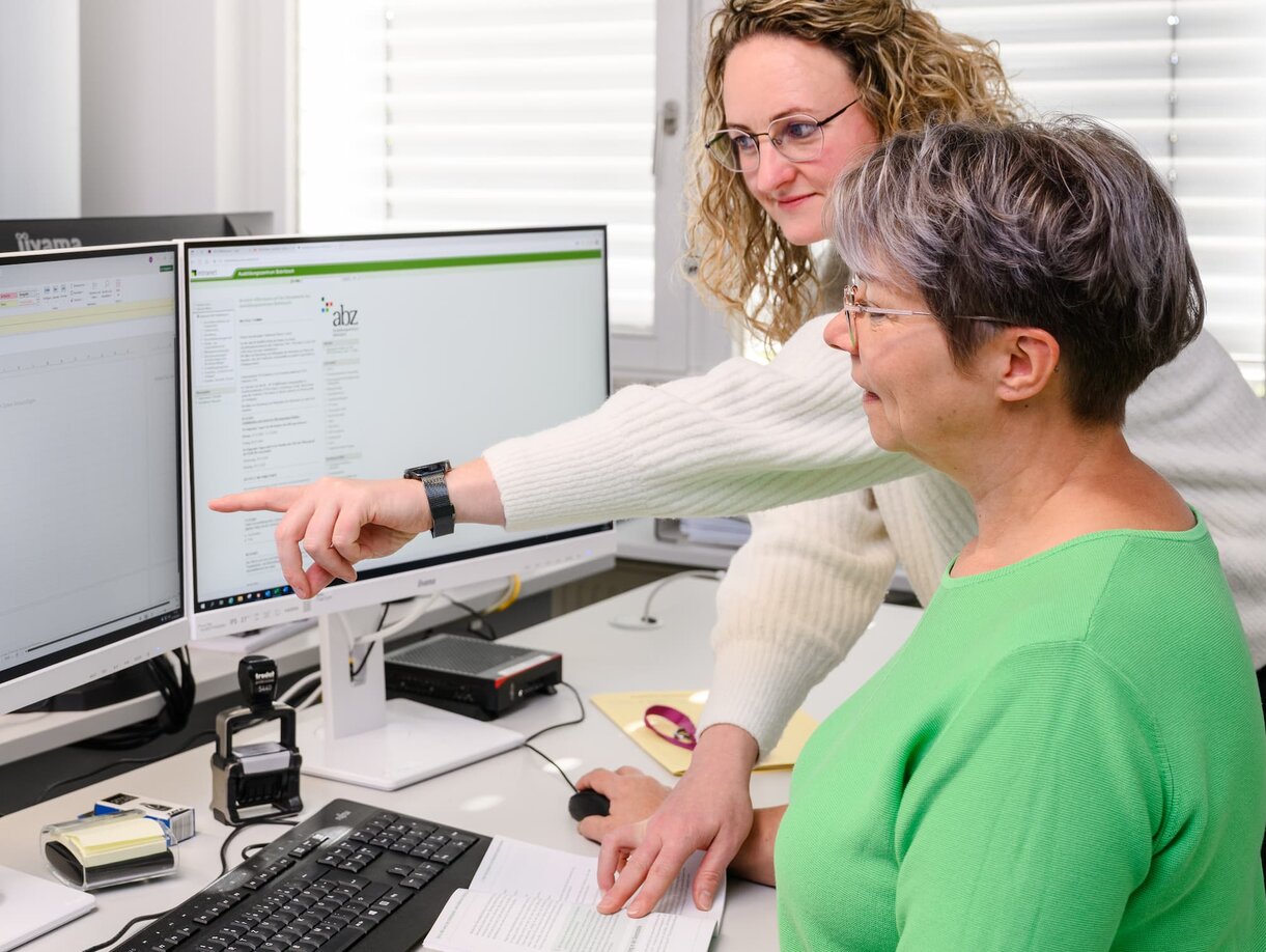 Zwei Sachbearbeiterinnen beraten sich an einem Schreibtisch; eine zeigt etwas auf einem der beiden Monitore