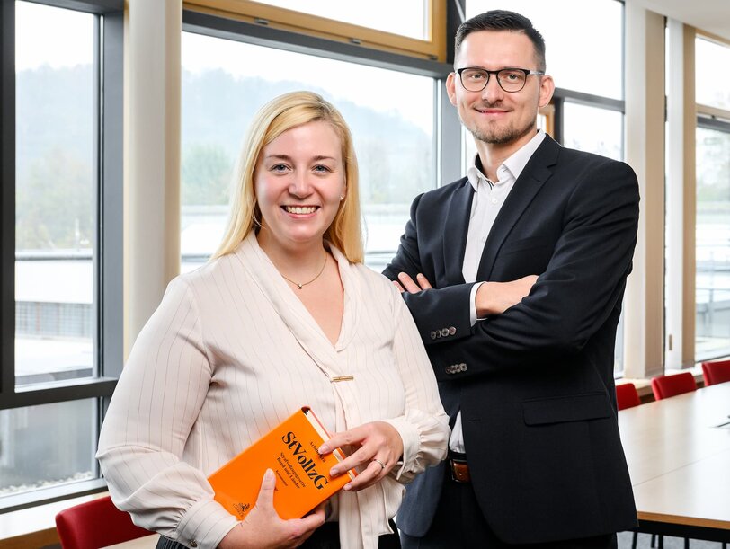 Eine Juristin und ein Jurist stehen in einem Raum des Justizvollzuges; die Juristin hält ein Buch unter dem Arm