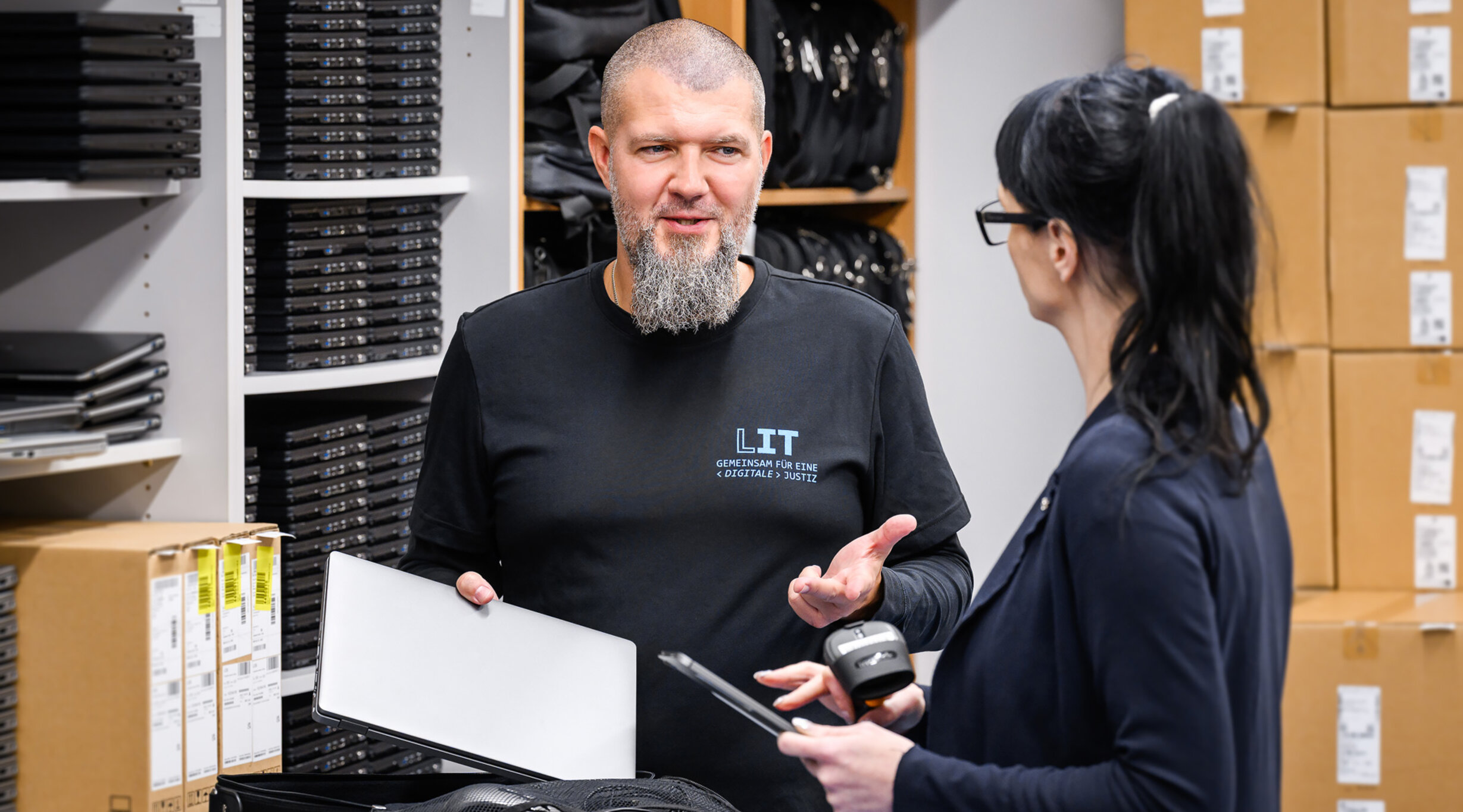 Ein Mitarbeiter und eine Mitarbeiterin der Leitstelle für IT für Beschaffung im Gespräch mit Notebooks im Lager