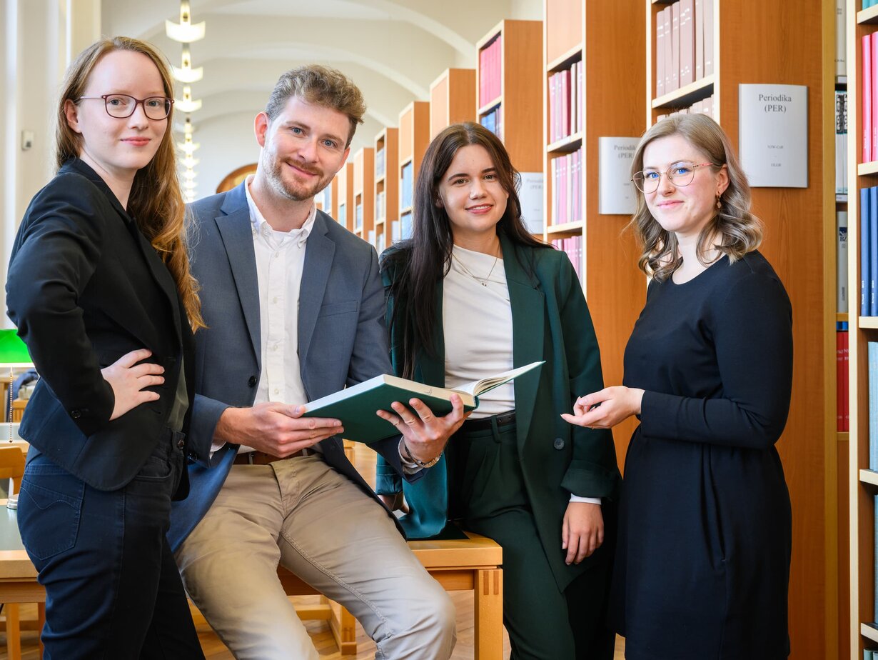 Eine Gruppe von Rechtspflegern in der Bibliothek; ein Rechtspfleger in der Mitte hält ein aufgeklapptes Buch