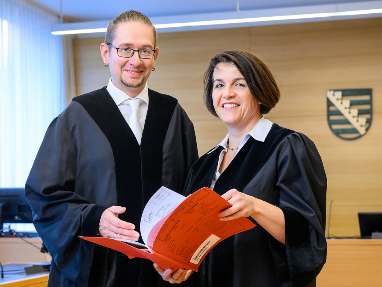 Ein Amtsanwalt und eine Amtsanwältin in einem Gerichtssaal und halten eine Akte geöffnet vor sich