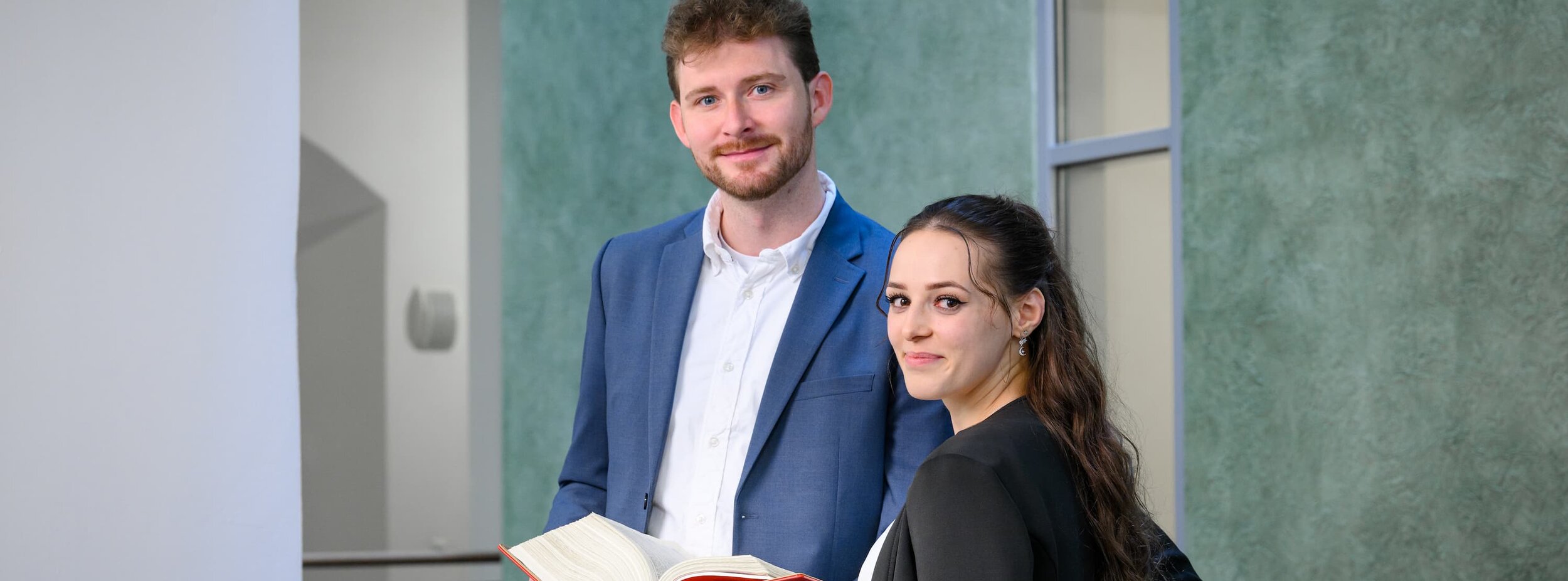 Ein Rechtspfleger und eine Rechtspflegerin stehen im Gang; der Rechtspfleger hält ein aufgeklapptes Buch